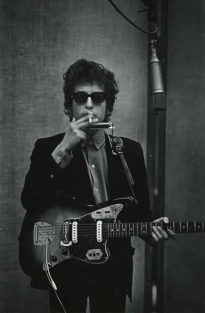 Bob Dylan in the studio with electric guitar, harmonica rack, and cigarette — black and white.